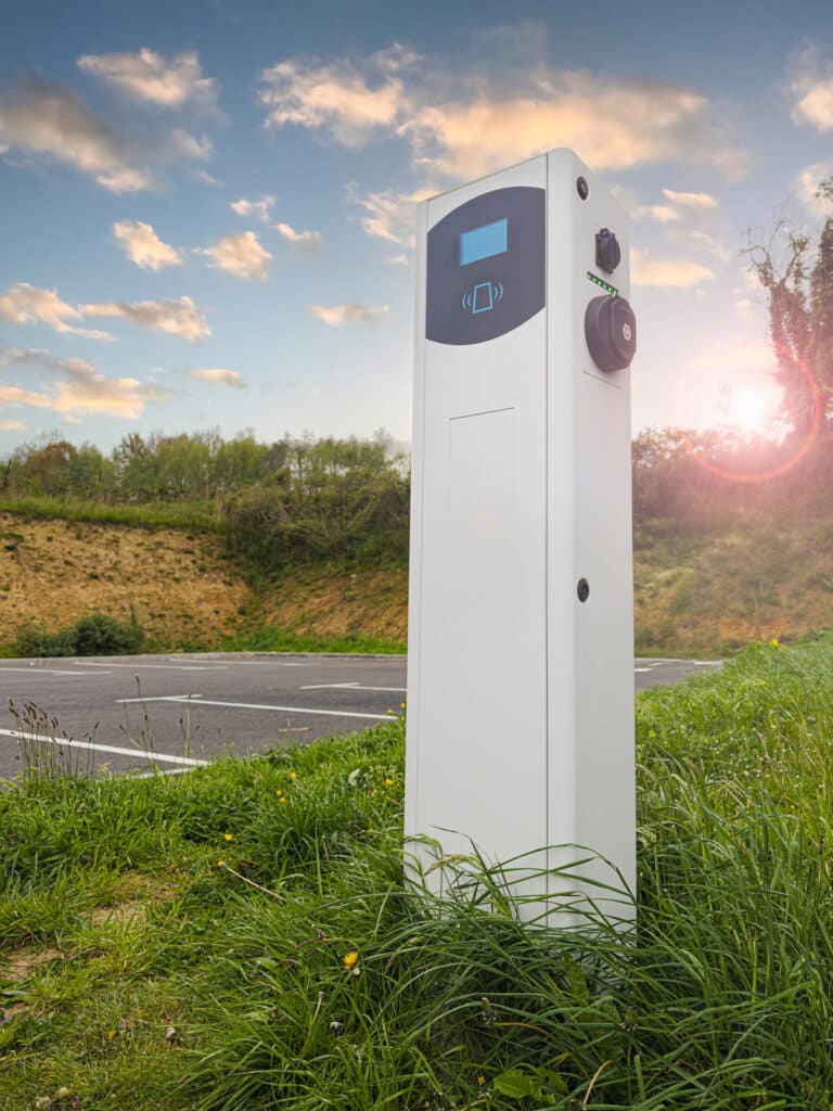 Borne de recharge VE moderne (écran bleu, symbole sans contact) dans l'herbe près d'un parking, sous un ciel nuageux et un soleil intense.