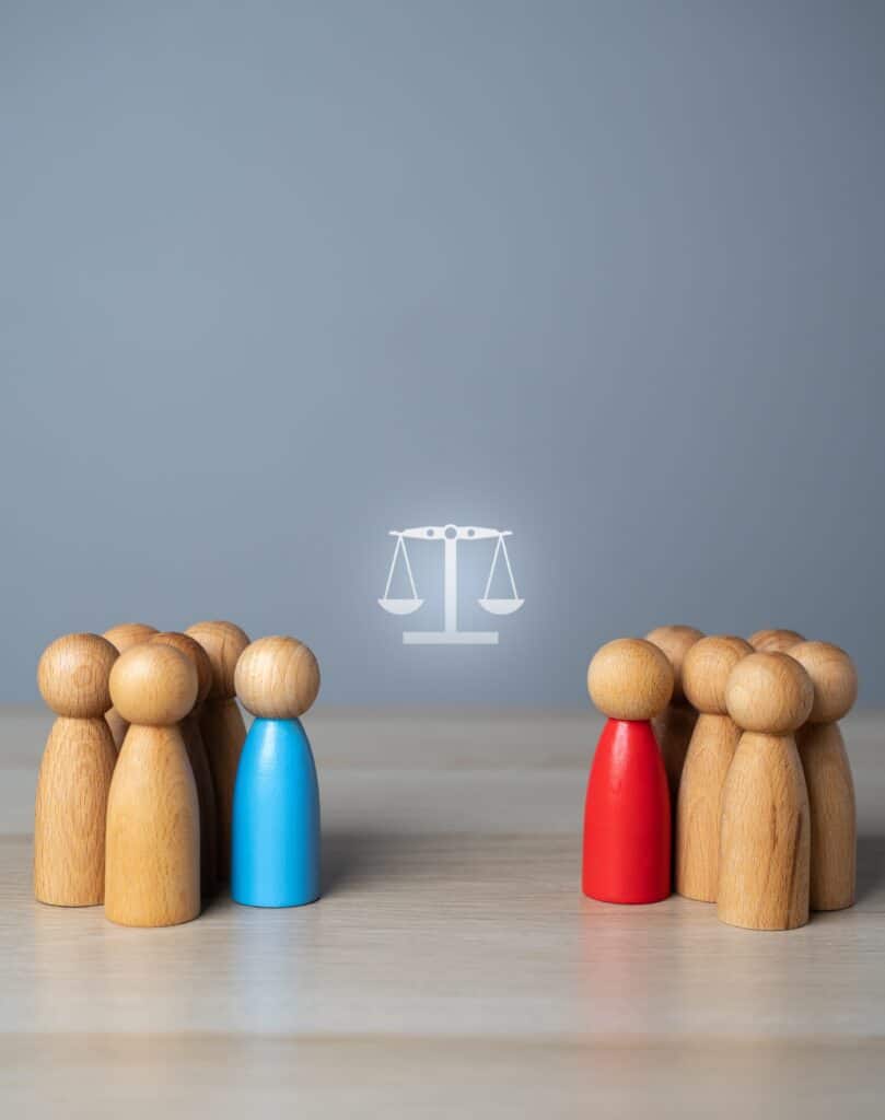 Deux groupes de figurines bois (leaders bleu et rouge) face à face, séparés par les Balances de la Justice. Symbole de litige.