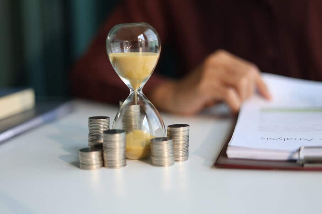 Temps, argent et analyse financière. Un sablier rempli de sable jaune au milieu de piles de pièces de monnaie sur un bureau, avec un document d'Analyse.